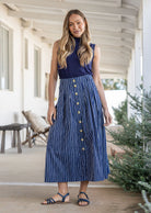 Woman wearing a navy basic top and button through shin length cotton skirt with box pleats, belt loops and pockets.