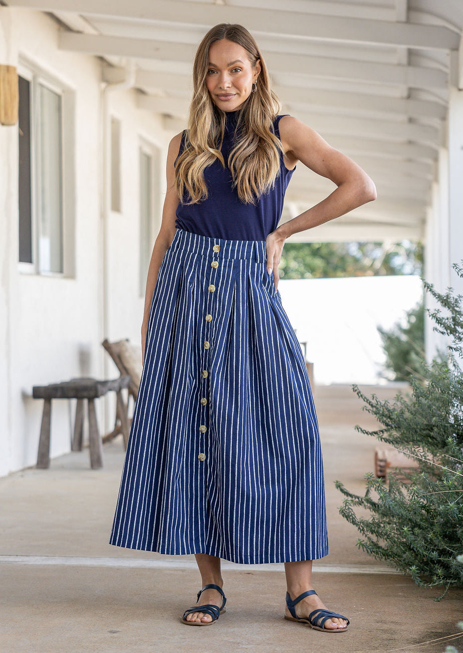 Woman stands wearing a navy top and striped print, shin length cotton skirt with button through front and box pleats.