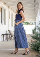 Woman stands sideways wearing a navy top and white striped on navy base print, shin length cotton skirt with button through front and box pleats.