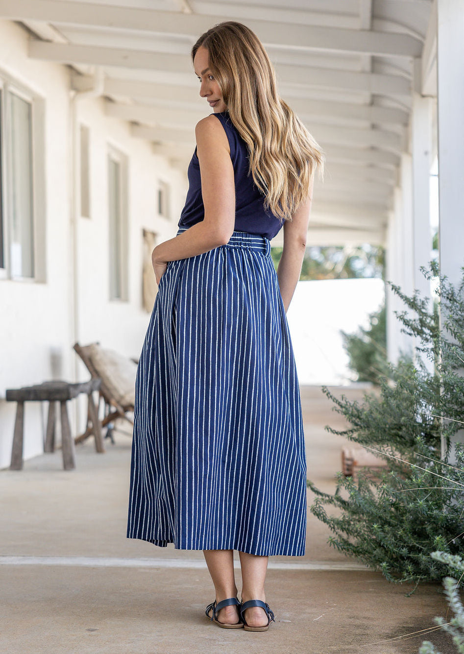 Woman stands backwards wearing a white striped on navy base print, shin length cotton skirt with button through front and box pleats.