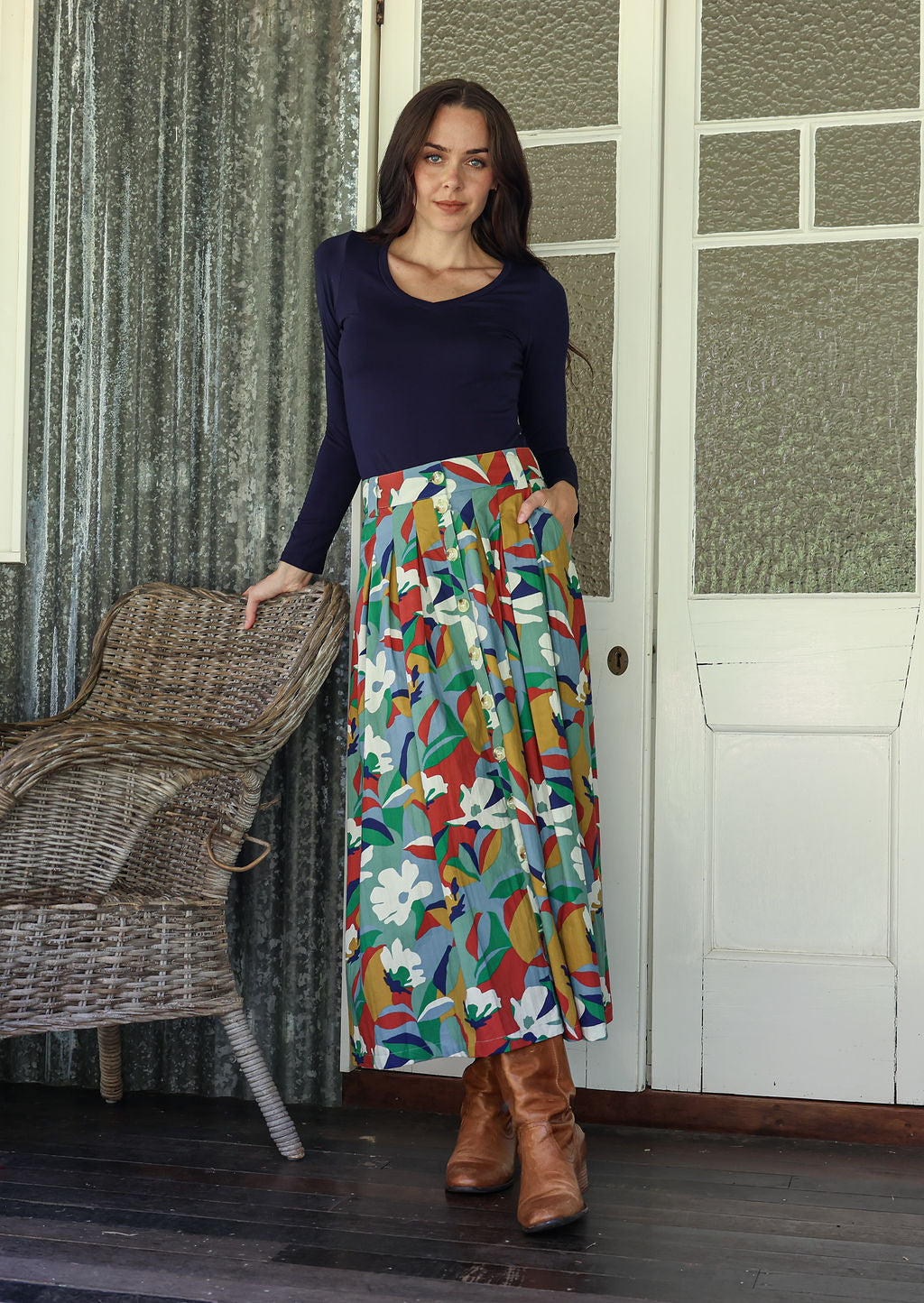 Woman wearing a colorful patterned skirt and brown boots standing in a room with a textured wall and wicker chair.
