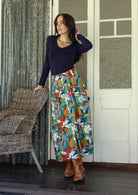Woman wearing a colorful skirt and navy top standing in a room with wicker chair and patterned walls.