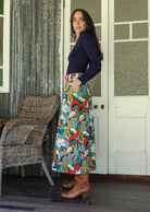 Woman stands sideways wearing a floral print, shin length cotton skirt with button through front and box pleats.