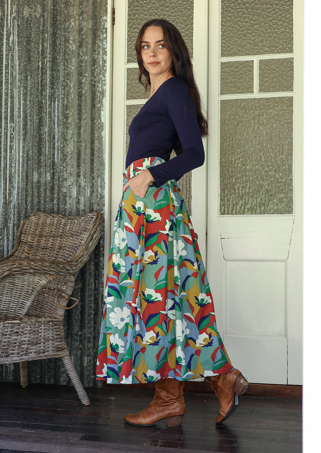 Woman wearing a colorful floral skirt and navy top standing in a room with wicker chair and patterned walls.