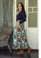 Woman wearing a colorful floral skirt and navy top standing in a room with wicker chair and patterned walls.