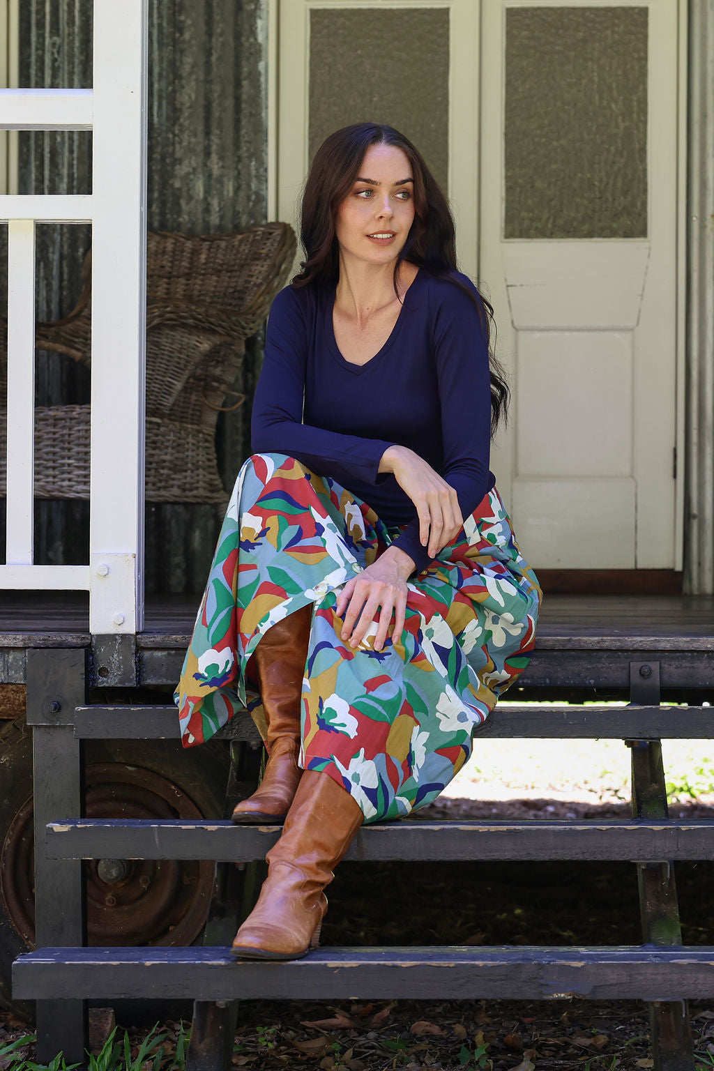 Woman sitting on a porch wearing a colorful skirt and brown boots.