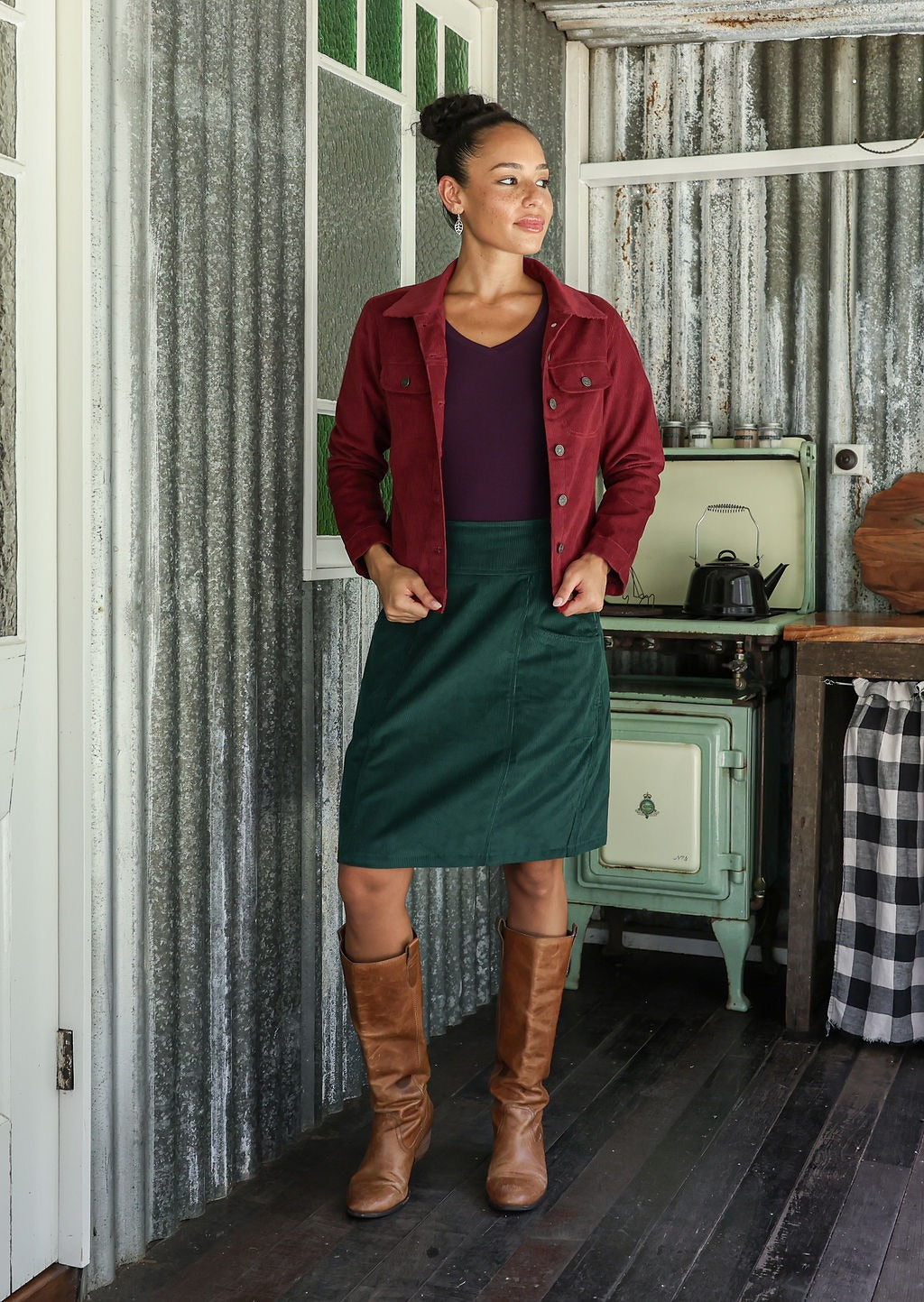 Woman in a deep, luxurious, and intense red-purple hue jacket and green corduroy skirt standing in a rustic room with a stove and checkered tablecloth.