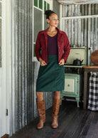 Woman in a deep, luxurious, and intense red-purple hue jacket and green corduroy skirt standing in a rustic room with a stove and checkered tablecloth.
