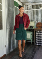 Woman wearing a corduroy jacket, green skirt, and brown boots standing in a rustic setting.