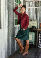 Woman in a corduroy jacket and green skirt standing in a rustic kitchen.