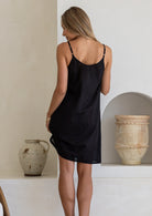 Black cotton slip undergarment with thin straps, shown on model from behind against plastered wall.