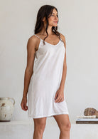 Model stands side on wearing white cotton slip undergarment with thin straps and lace hem, above knee length
