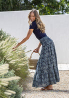 Woman in a floral skirt and navy top standing outdoors with plants and a white wall in the background