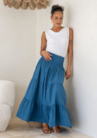 Woman wearing a white singlet top and blue tiered cotton maxi skirt with a wide shirred waistband, leaning against a wall