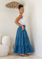 Woman wearing a blue tiered cotton maxi skirt with a wide shirred waistband, paired with a white singlet top