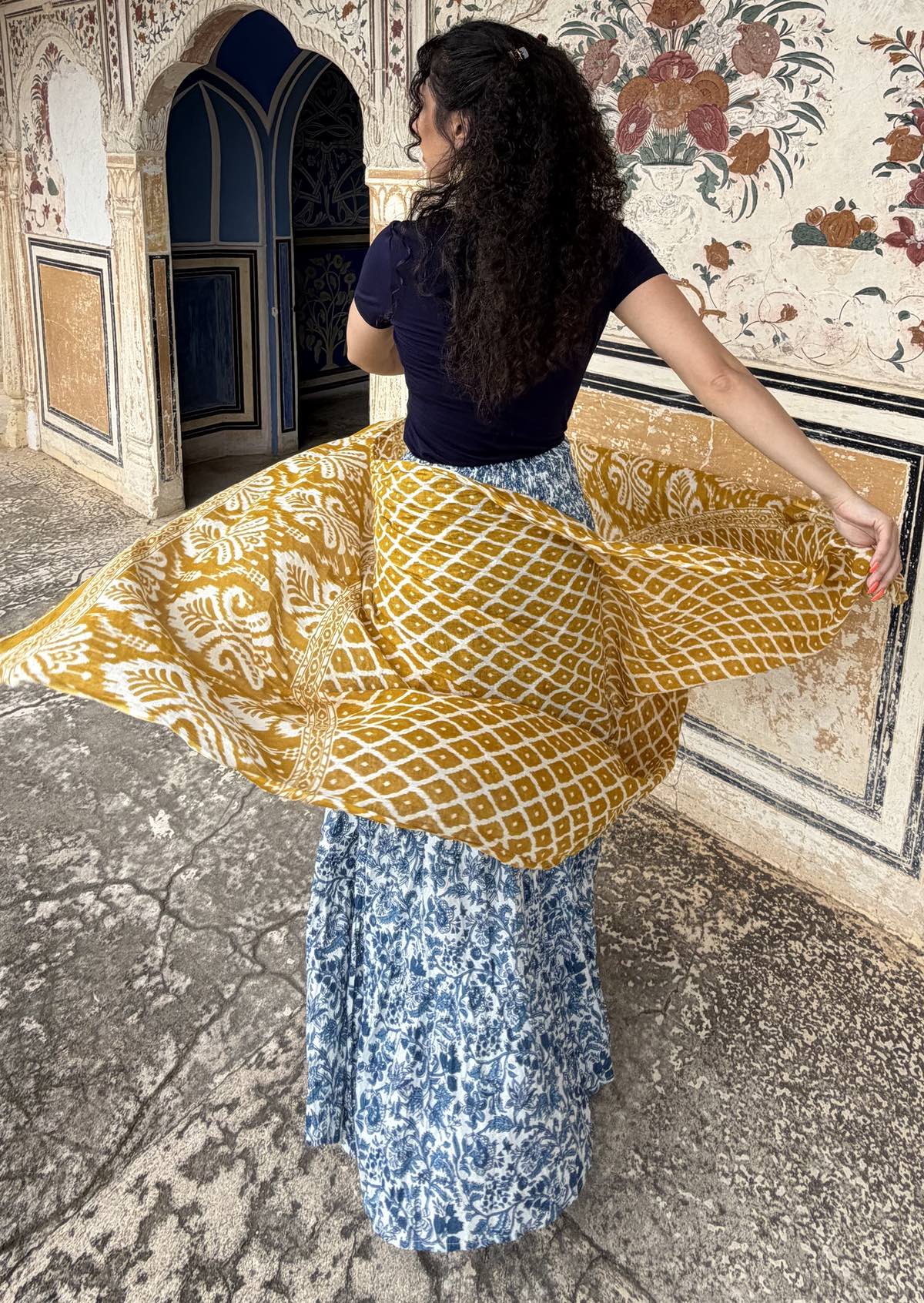 Woman twirls wearing a white based blue floral cotton maxi skirt paired with a navy blue stretch t-shirt, swirling a yellow shawl around her