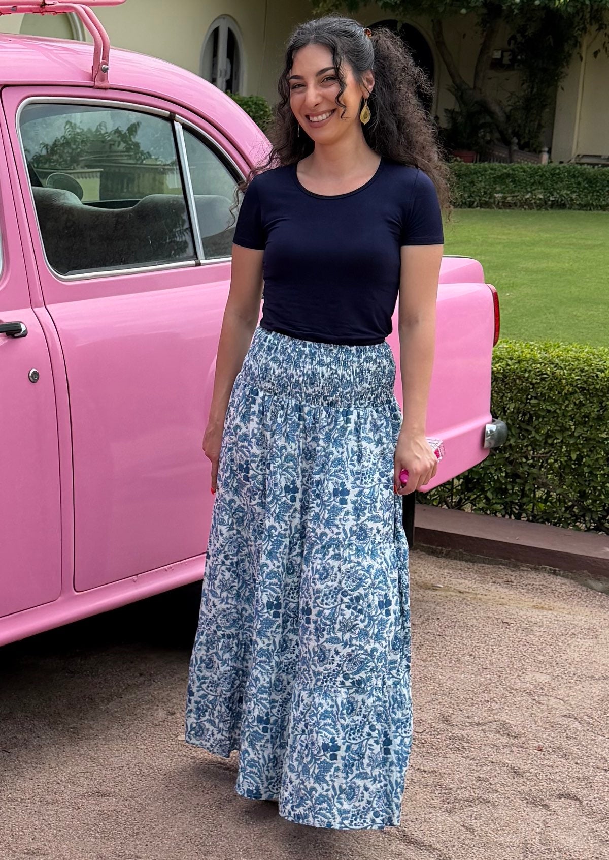 Woman wears a white based blue floral cotton maxi skirt paired with a navy blue stretch t-shirt, standing in front of a pink car