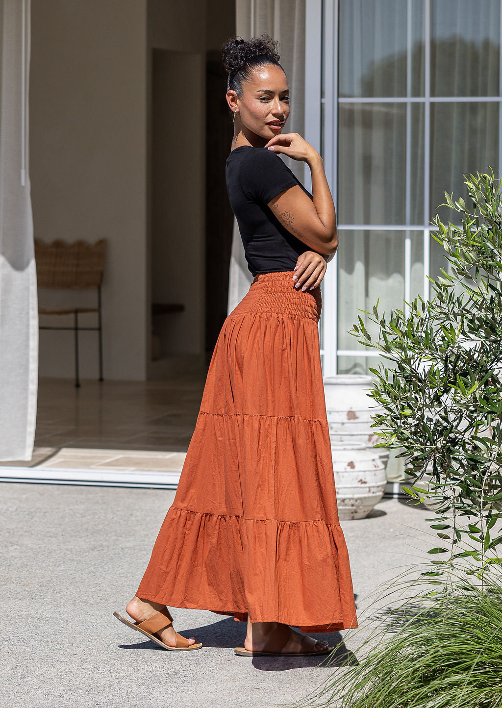 Woman standing side on in a rust coloured cotton maxi skirt with a wide elasticated waistband, paired with a black stretch t-shirt