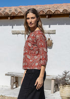 Woman stands side on in an earthy red based floral cotton top with 3/4 sleeves