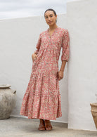 Woman poses in rose pink based floral cotton maxi dress with buttoned bodice, 3/4 sleeves and flowy three tiered maxi skirt