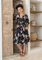 Model wears a black based floral printed cotton wrap dress with V-neckline, 3/4 sleeves, a wide waist-tie and pockets