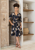 Model wears a black based floral printed cotton, over the knee length wrap dress with V-neckline, 3/4 sleeves, and a wide waist-tie