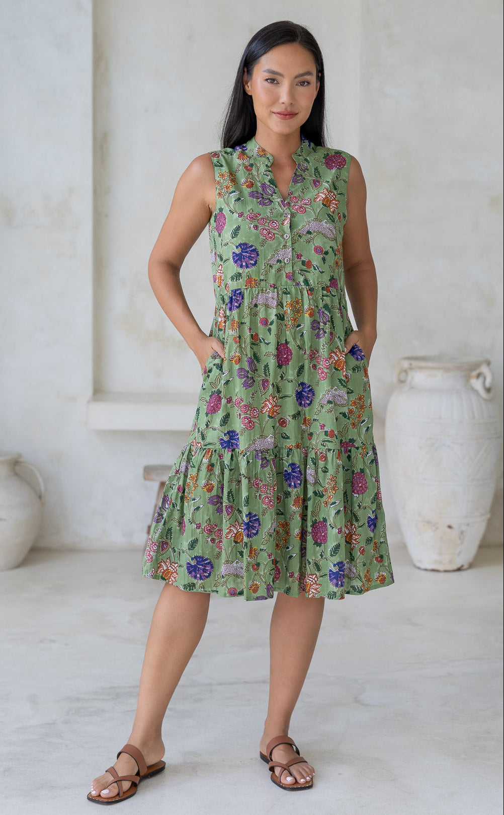 Woman wearing a moss green floral sleeveless cotton dress with a buttoned bodice, mandarin collar and hands in pockets.