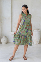 Woman holding skirt out to the side of a green floral sleeveless, knee length dress with a buttoned bodice, tiered skirt and pockets. 