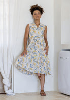 Woman wearing a knee length cotton sleeveless dress with buttoned bodice and  two tiered skirt in neutral based floral print.