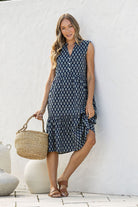 Woman in a blue patterned dress holding a woven basket against a white wall.