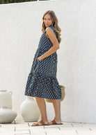Woman stands sideways wearing a sleeveless navy blue based floral cotton dress with pockets, mandarin collar and a two tiered skirt.