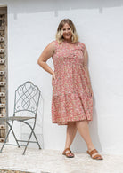 Woman stands wearing a sleeveless rose pink floral cotton dress with pockets, mandarin collar and a two tiered skirt.
