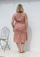 Woman stands backwards wearing a sleeveless rose pink floral cotton dress with pockets, mandarin collar and a two tiered skirt.