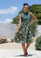 Woman holding skirt out to the side of a green floral sleeveless, knee length dress with a buttoned bodice, tiered skirt and pockets. 