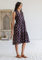 Woman stands sideways wearing a sleeveless black floral cotton dress with pockets, mandarin collar and a two tiered skirt.