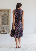 Woman stands backwards wearing a sleeveless navy floral cotton dress with pockets, mandarin collar and a two tiered skirt.