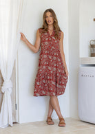 Woman wearing a knee length cotton sleeveless dress with buttoned bodice and  two tiered skirt in red based floral print.