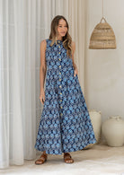 Woman wearing a navy based floral cotton sleeveless dress standing in a room with white curtains.