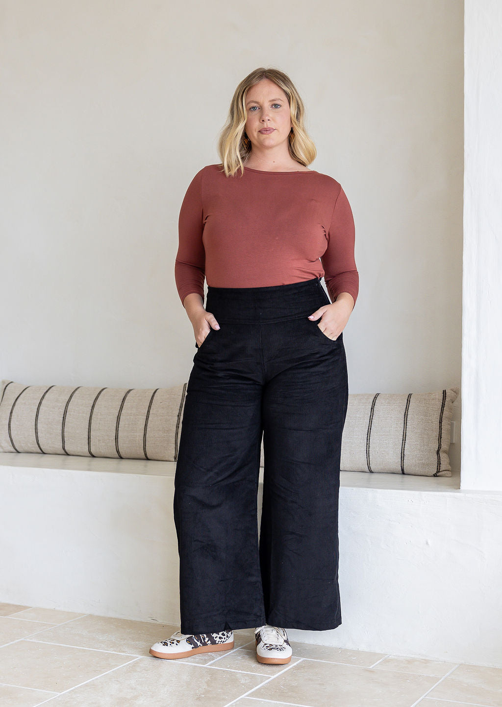 Woman standing wearing black cotton corduroy wide leg pants and a dusty pink top with both hand in pockets 