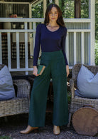 Woman wearing a navy blue top and green corduroy pants standing on a porch with wicker furniture and cushions.