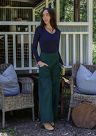 Woman in a navy top and green pants standing on a porch with one hand in pocket