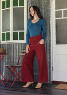 Woman in blue long sleeve top and red cotton corduroy pants standing in a room with a textured wall and window.