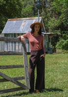 Woman wearing a brown pants standing by a wooden fence in a rural setting with a rustic building in the background.