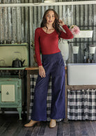 Woman in red long sleeve top and blue cotton corduroy wide leg pants standing in a rustic kitchen.