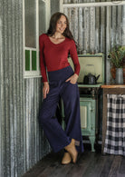 Woman in red long sleeve top and blue cotton corduroy pants standing in a rustic kitchen with hand in pocket