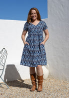 Model wearing a cotton knee length dress with empire waist, short sleeves and hand in pockets paired with brown boots. 