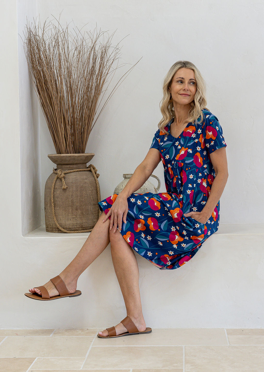 A woman wearing a short-sleeve navy dress with a colorful floral print sits on a white ledge in a sunlit room.
