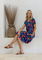 A woman wearing a short-sleeve navy dress with a colorful floral print sits on a white ledge in a sunlit room.
