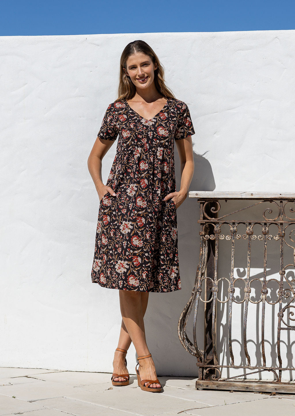 Woman wearing a floral dress standing against a white wall with a metal gate.