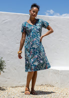 Woman wearing a teal cotton floral dress standing against a white wall with blue sky.